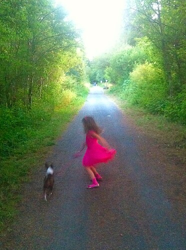 A child in a pink dress appears to be twirling or running playfully with a dog along a tree-lined path. (Captioned by AI)