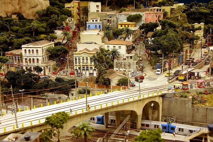 Dies ist eine malerische Miniaturansicht einer Stadt mit farbenfrohen Gebäuden, Palmen, kleinen Fahrzeugen und einem Zug, der unter einer Brücke fährt, die alles zusammenhält <code>. (Captioned by AI)