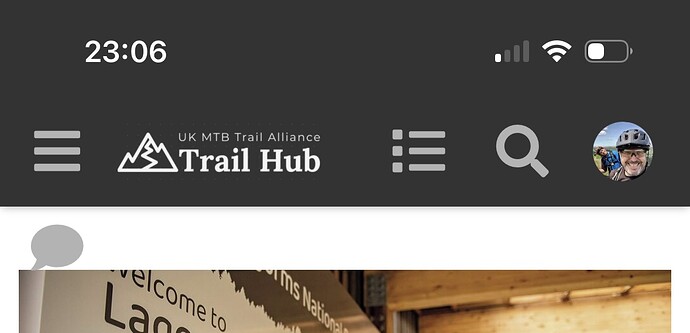 The image shows the header section of a mobile app called "Trail Hub" by the UK MTB Trail Alliance, featuring their logo, menu icons, and a profile picture, along with a blurred image in the background that seems to be of a "Welcome to..." sign, possibly for a trail or outdoor area. (Captioned by AI)