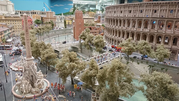 The image displays a detailed miniature model of an urban scene with scale replicas of buildings, vehicles, and people, featuring the Colosseum in the background. (Captioned by AI)