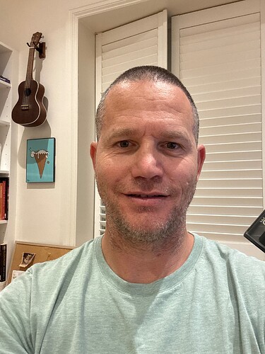 Un hombre con una camiseta verde claro está de pie en el interior con una sonrisa, cerca de una ventana con persianas y una guitarra en la pared. (Subtitulado por IA)