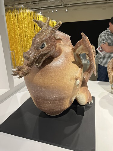 يُظهر الرسم التوضيحي منحوتة كبيرة ذات ملمس تشبه تنينًا يخرج من وعاء، معروضة في معرض فني مع تركيب يشبه الذهب والزينة في الخلفية وشخص يراقب العمل الفني إلى اليمين. (تم التعليق بواسطة الذكاء الاصطناعي)