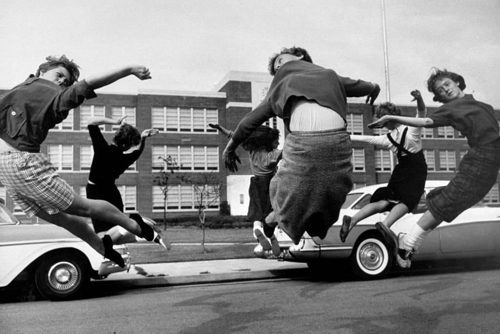 A group of young people appear to be playfully jumping off the bumper of a parked car, evoking a sense of carefree youth in a bygone era. (Captioned by AI)